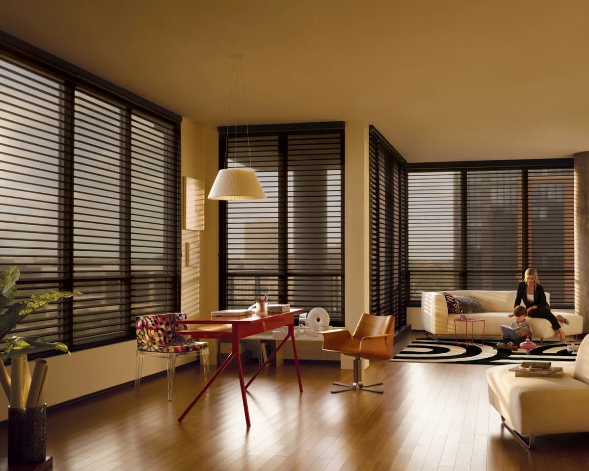 Sunlit living room with sheer horizontal shades across floor-to-ceiling windows, a woman and child reading on the sofa