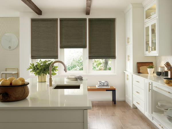 Woven Roman shades in a white farmhouse kitchen, showing natural texture and light filtering of woven wood material over the sink area